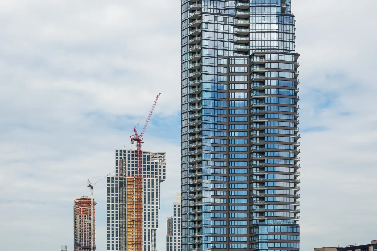 The Greenpoint tower at 21 India Street viewed from across the East River, showing its full glass facade and the KAWS sculpture at its base